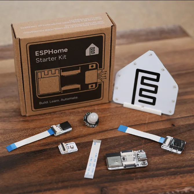 Kit ESPHome: Empieza Hoy 3 La caja del Kit de Inicio ESPHome junto a su contenido desempaquetado: módulo de notificación, placa principal, sensor de movimiento, módulo de botón, cables planos y soporte, dispuestos sobre una superficie de madera.